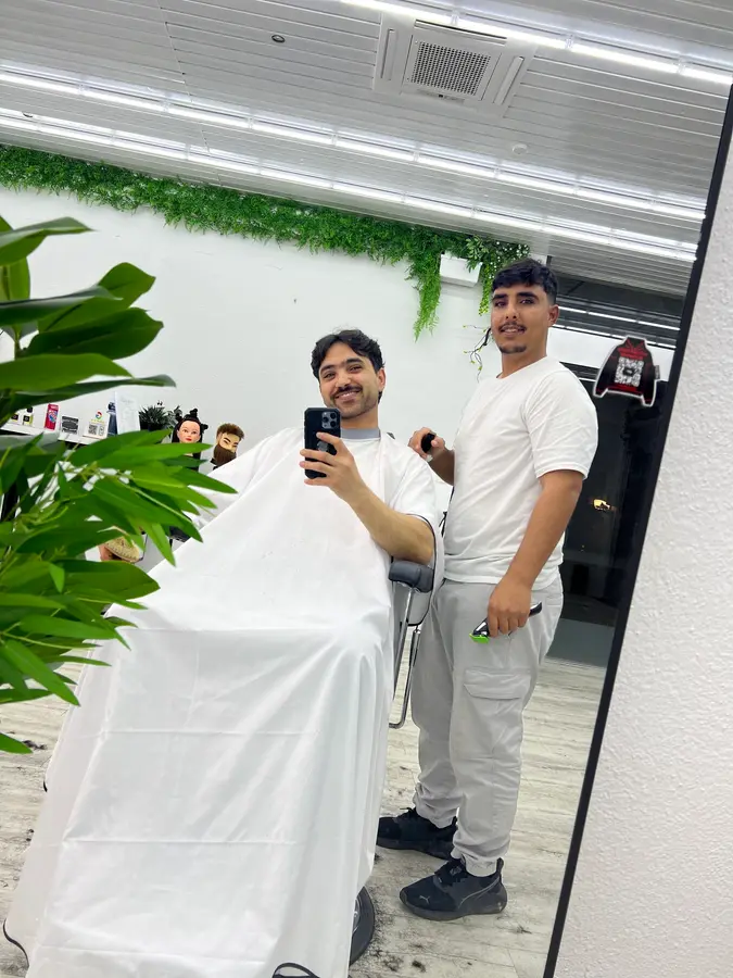 Blick in den hellen Salon mit grüner Pflanzenwand: ein Barbier neben einem Kunden im Umhang